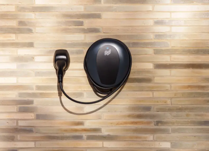Tethered home charger on a light brick wall, with the cable and connector stored beside it